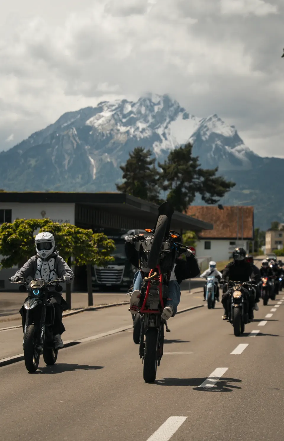 Motorradfahrer macht einen Wheelie, dahinter weitere Biker und ein verschneiter Berg